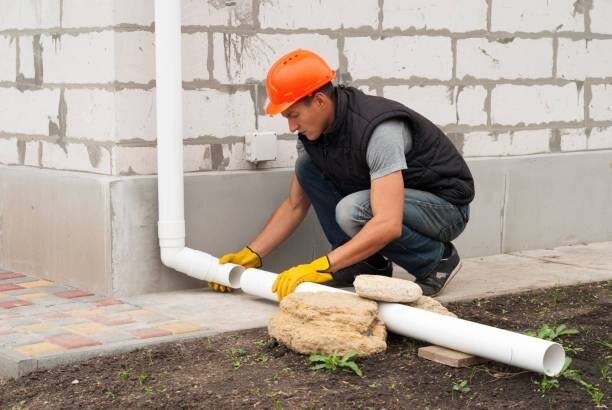 Worker fitting a white downspout extension along the ground at the base of a wall